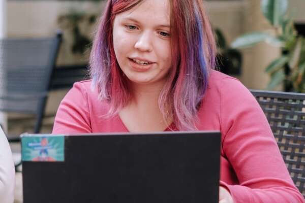 A woman with pink hair and a pink top is sitting in front of a laptop.