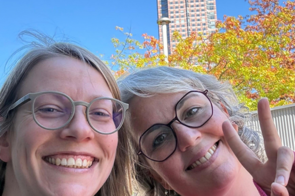 Angie and Helen are smiling in the camera, in the background is the Frankfurt Fair tower in front of the blue sky.