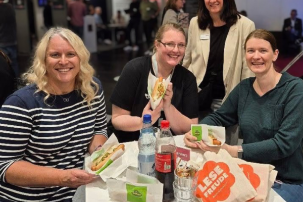 Four women sit at a desk and eat MVB-hotdogs.