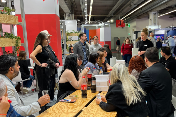 People are sitting around a table at the MVB-booth and listeing to a women dressed in black.