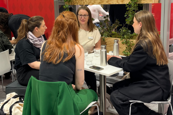 Four young women sit at the desks of MVB-booth and chat.