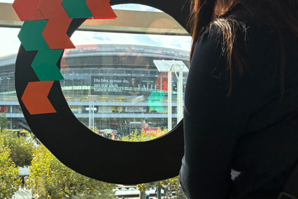A woman helds a handcrafted VLB-magnifying glass in front of a window and points to the building of hall 3 at Frankfurt Book Fair.